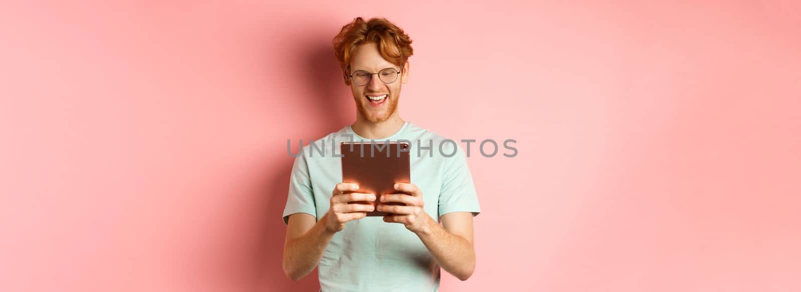 Happy guy with red hair and beard using digital tablet, reading screen and smiling, standing over pink background.