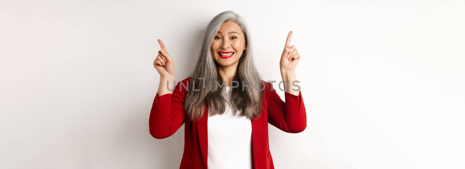 Happy mature woman in red blazer and makeup, smiling and showing advertisement on top, pointing fingers up at logo, white background by Benzoix