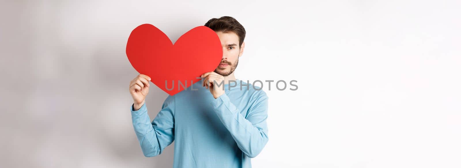 Young caucasian man holding Valentines day romantic heart, cover half of face with cutout and smiling, searching for love date, white background.