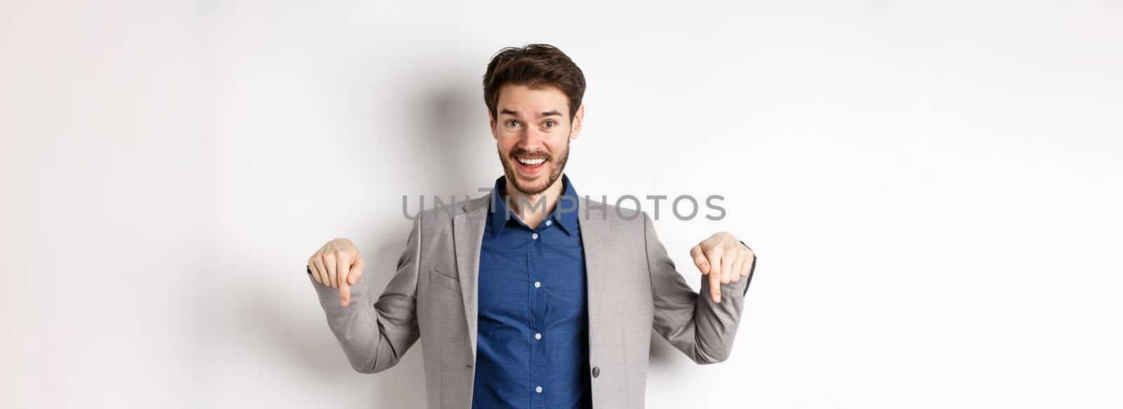 Hey check this out. Happy businessman in suit pointing fingers down and smiling, inviting to event, showing link, standing on white background.