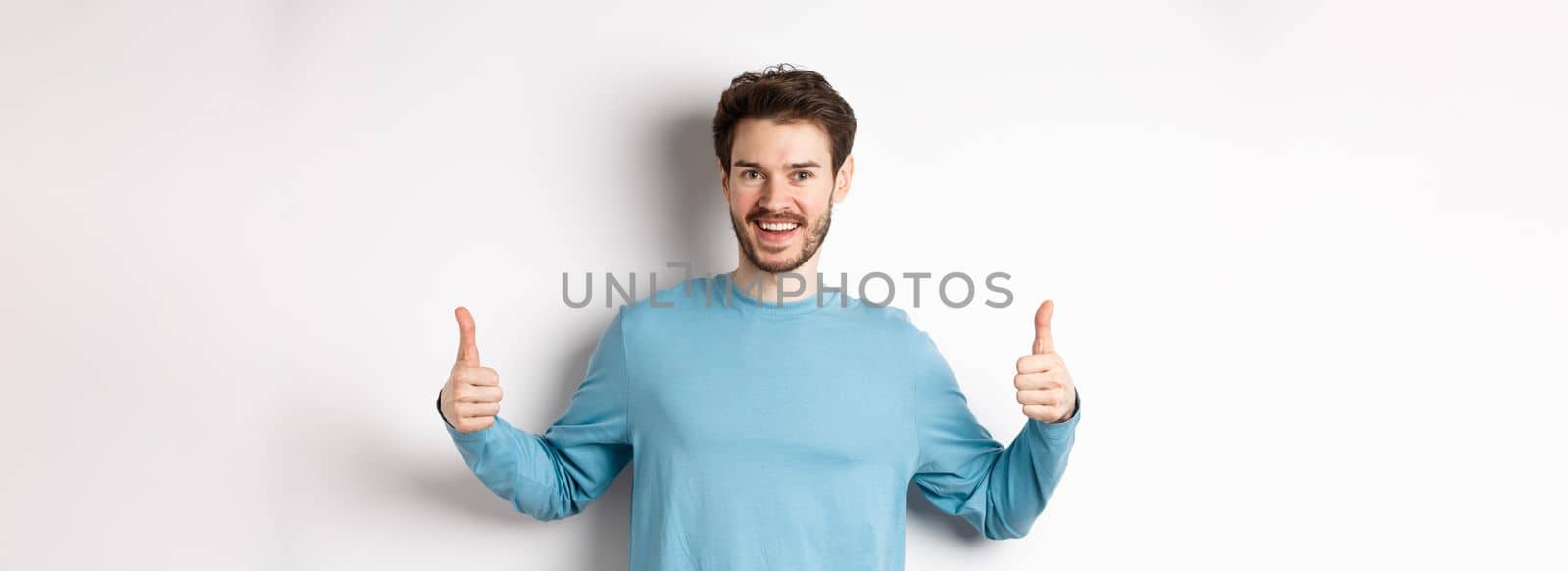 Cheerful young man showing thumbs up and smiling, recommend good product, praising choice, standing on white background.