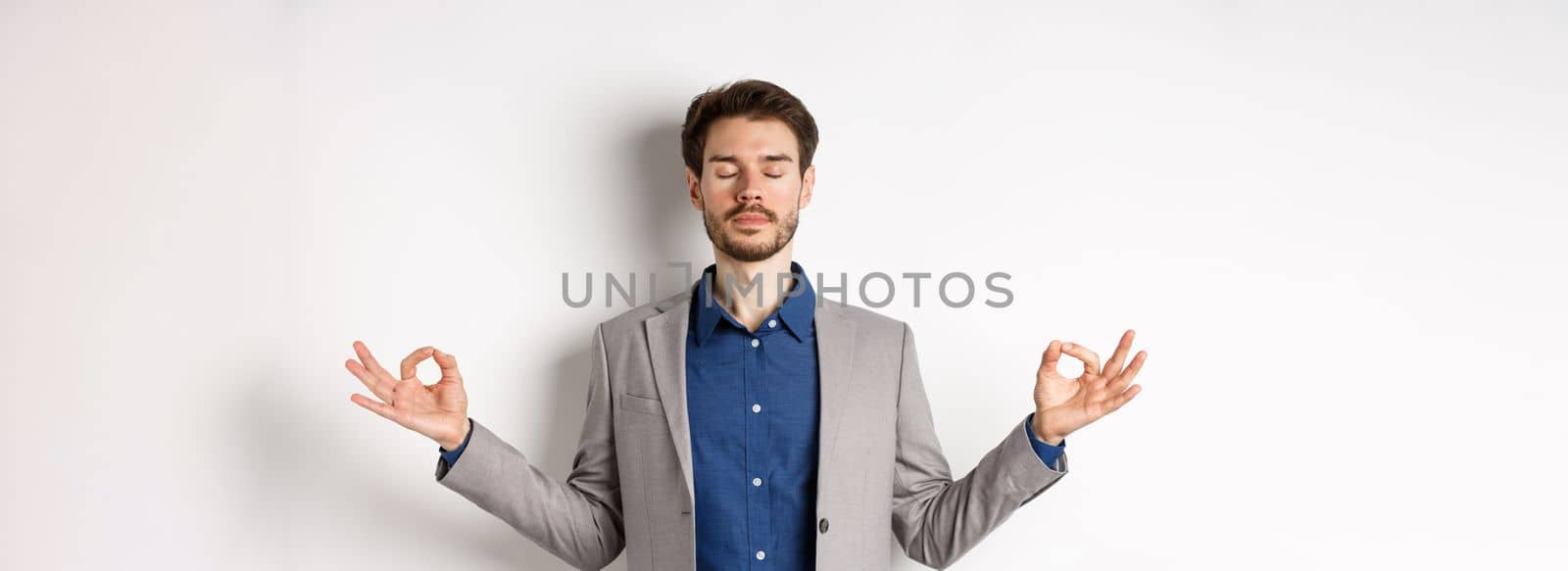 Calm and focused businessman meditating with eyes closed and hands spread sideways, finding peace in meditation, practice yoga breathing, standing on white background by Benzoix