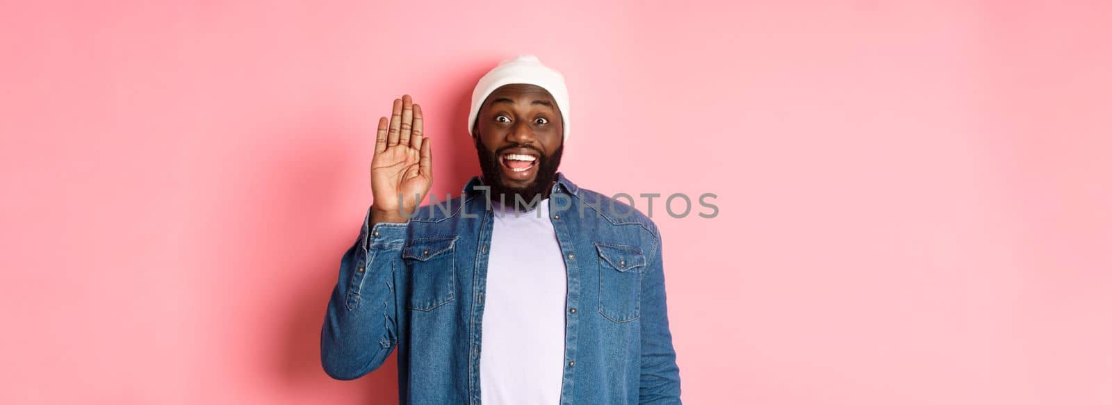 Friendly smiling Black man saying hello, waving hand, greeting you, standing over pink background by Benzoix