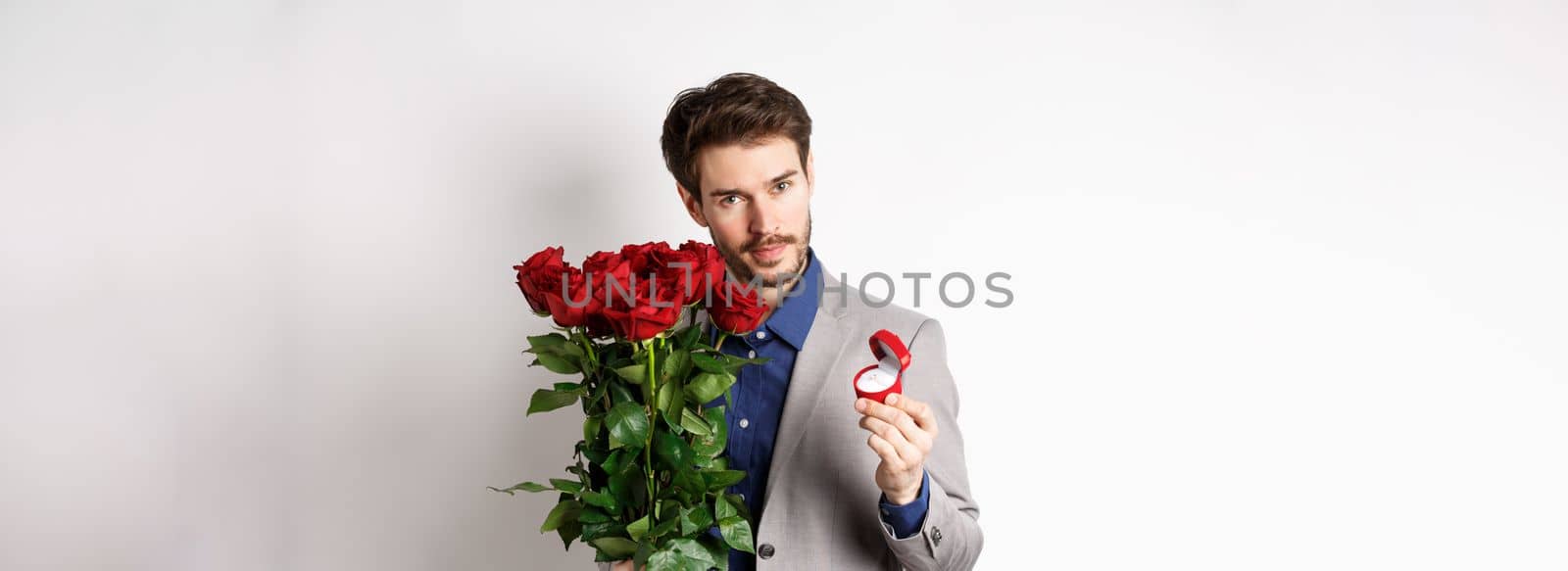 Handsome man in suit, showing engagement ring and looking romantic at camera, standing with red roses over white background. Valentines day and love concept by Benzoix