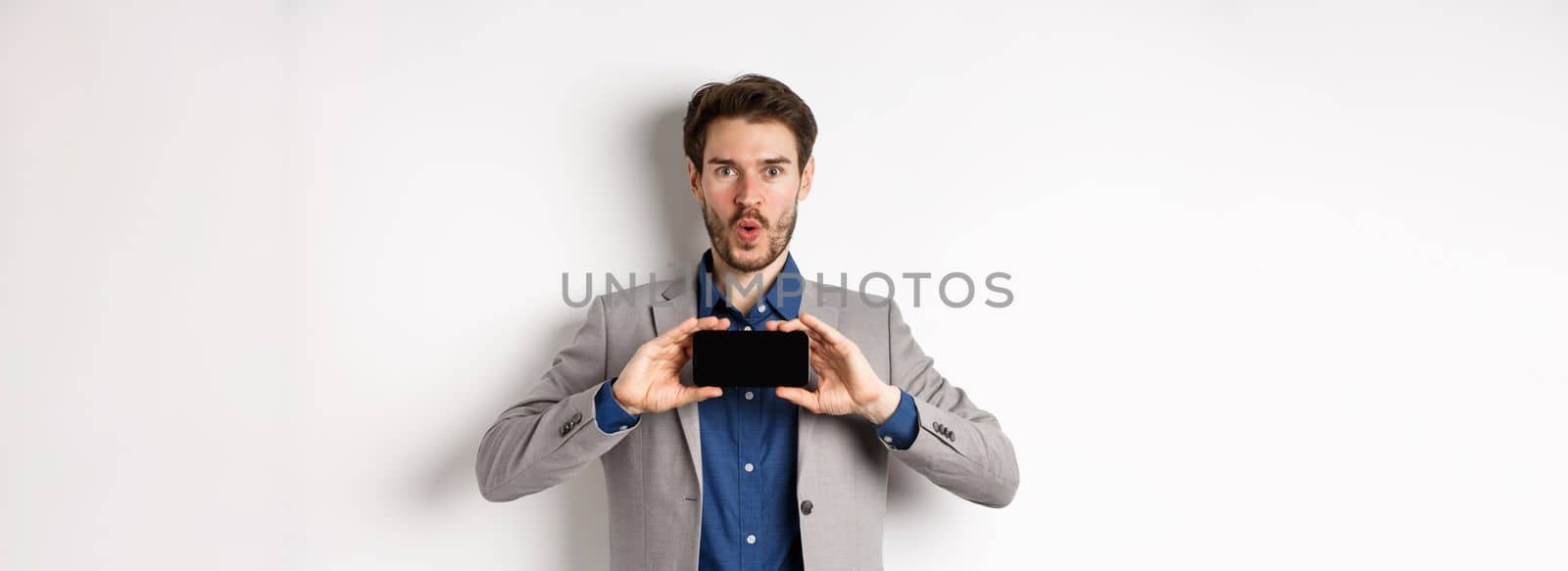 E-commerce and online shopping concept. Excited young man in business suit show empty smartphone screen and say wow amazed, white background.