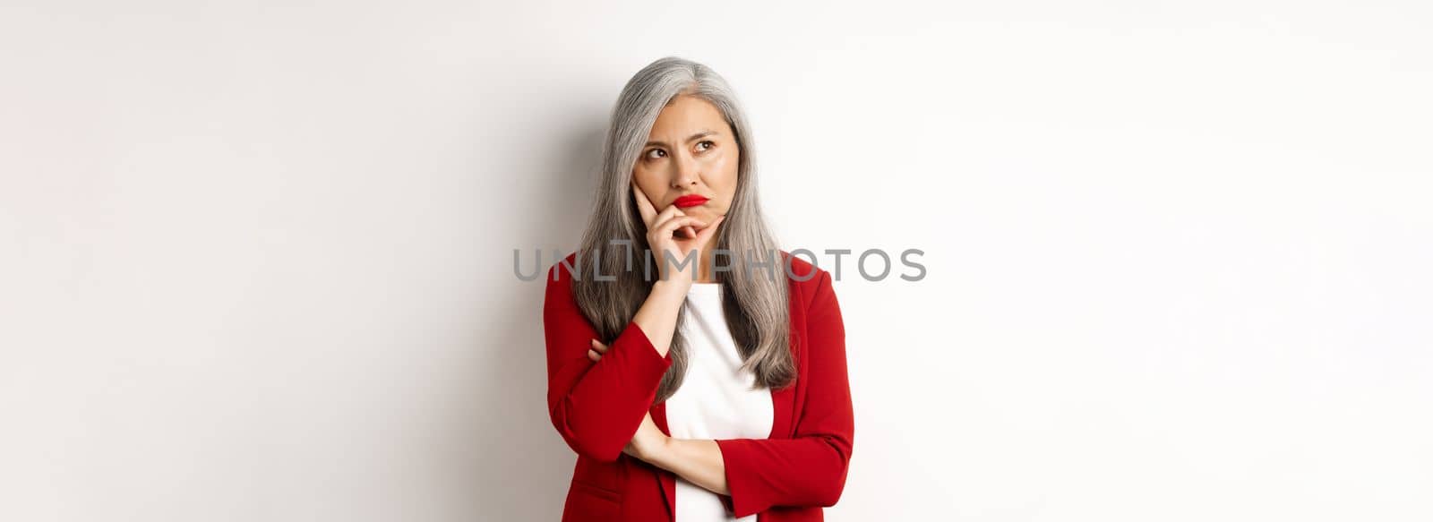 Troubled asian businesswoman in red blazer and lips, pouting and looking left annoyed, standing against white background.