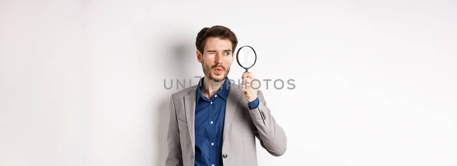 Handsome male model in suit looking through magnifying glass with interest, seeing something aside, white background.