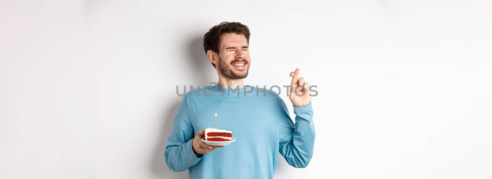 Celebration and holidays concept. Young man celebrating birthday, cross fingers for good luck, making wish on bday cake with lit candle, standing over white background.
