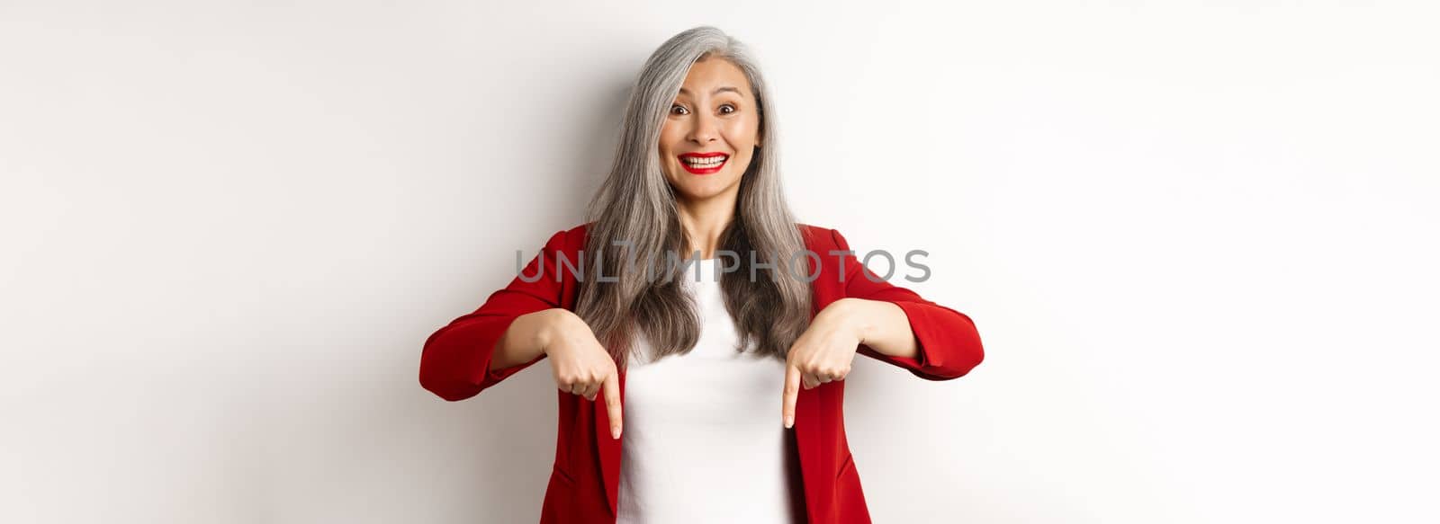 Cheerful asian female office worker pointing fingers down, smiling happy, showing promo offer, white background.