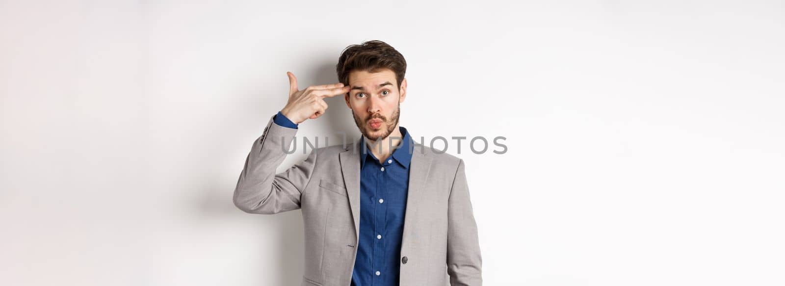 Annoyed guy in business suit shoot himself with hand gun near head, look distressed and tired after work, standing on white background.
