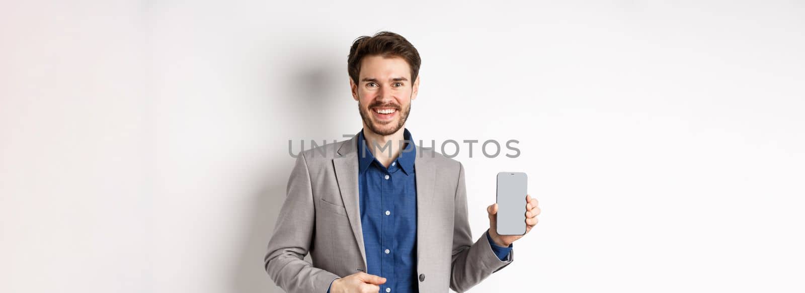 Handsome smiling salesman in suit showing empty smartphone screen, demonstrate an app, standing on white background.