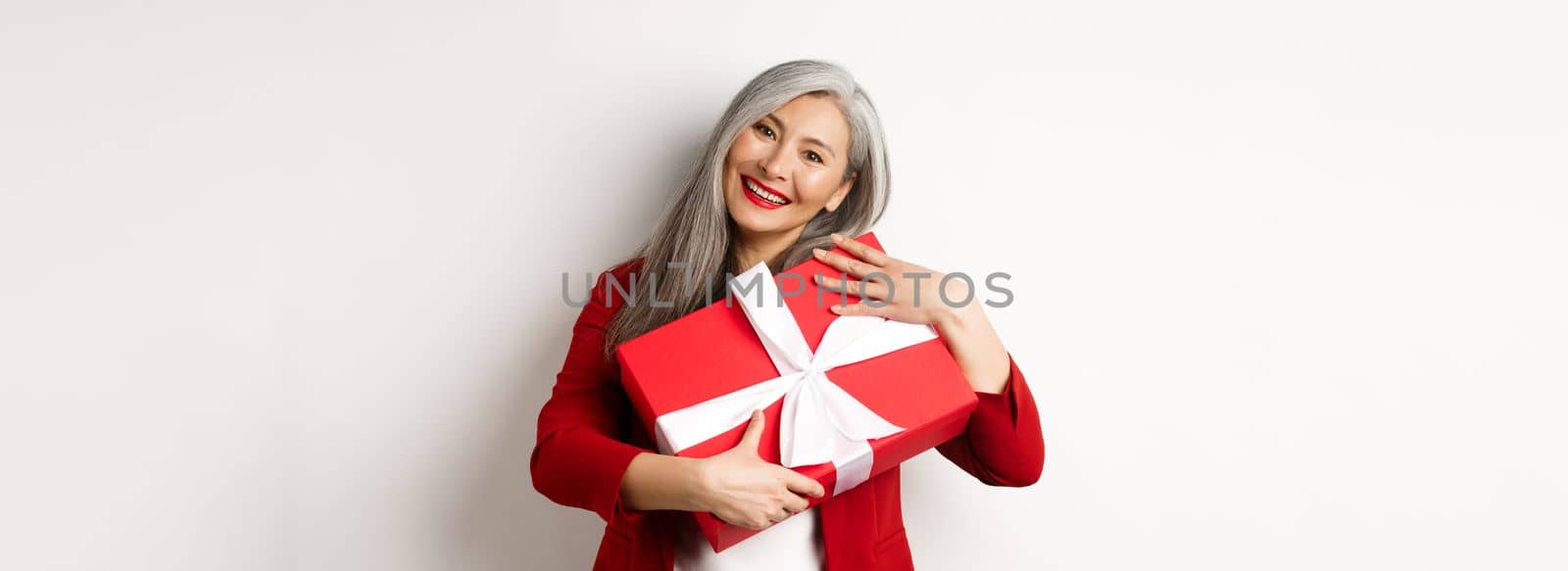 Happy asian grandmother hugging red gift box and smiling grateful, thanking for present, standing over white background.