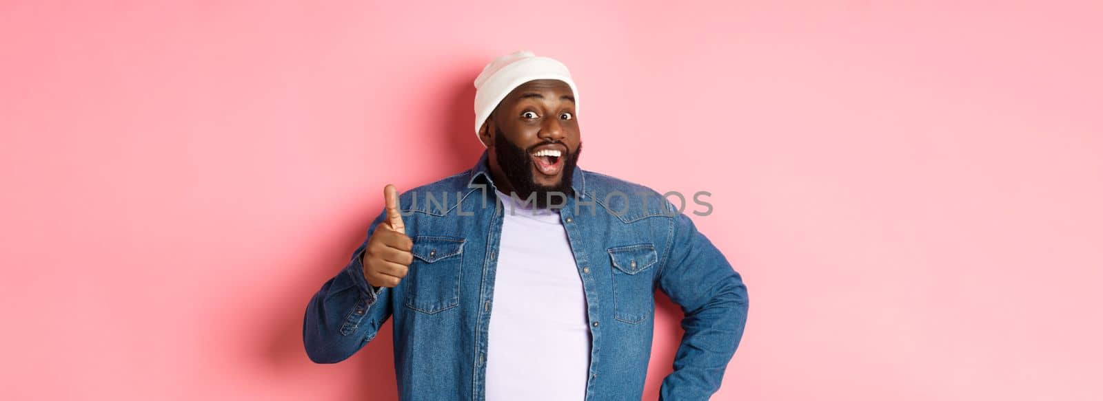 Happy african-american male model showing thumb-up and saying yesm smiling satisfied, standing over pink background.