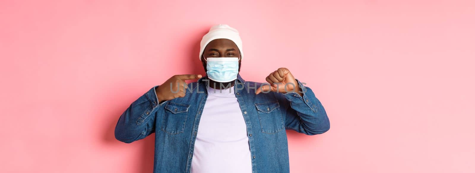 Coronavirus, lifestyle and global pandemic concept. Displeased hipster guy pointing at face mask, showing thumbs-down, dislike wearing it, pink background.