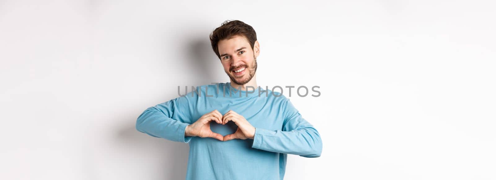 Handsome boyfriend saying I love you, showing heart gesture and smiling at camera, express love and romantic feeling, standing over white background by Benzoix