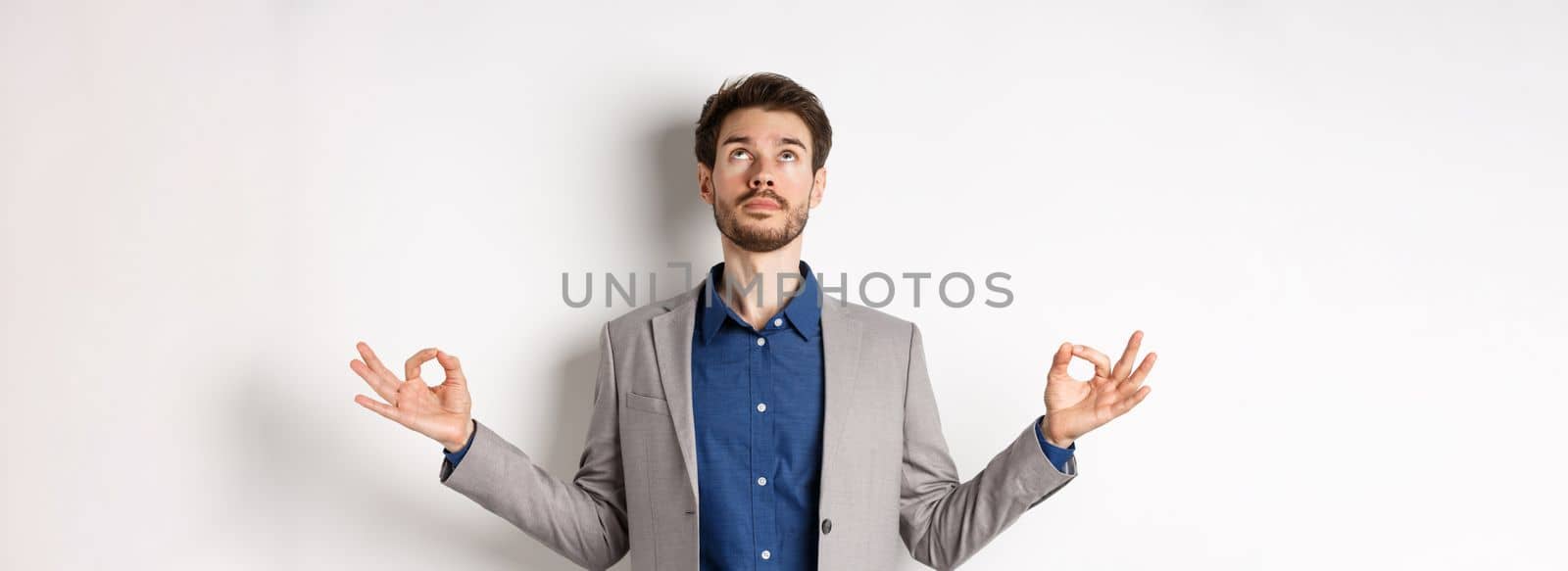Calm ceo manager in suit relaxing with meditation, looking up and holding hands in zen mudra sign, standing on white background.