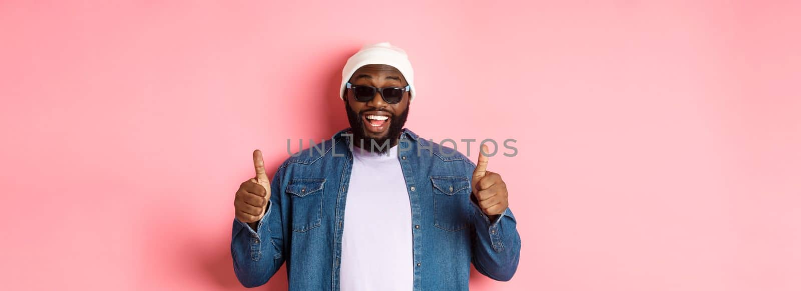 Satisfied african-american hipster guy in beanie and sunglasses showing thumbs-up, approve and praise good choice, smiling pleased, pink background.
