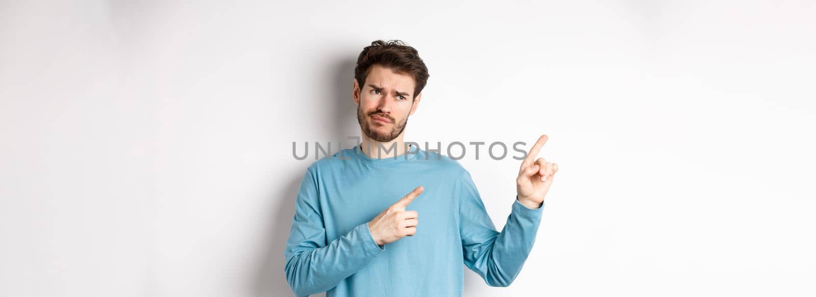 Skeptical and unimpressed young man grimacing, pointing fingers and looking at bad promotion, standing dissatisfied over white background.