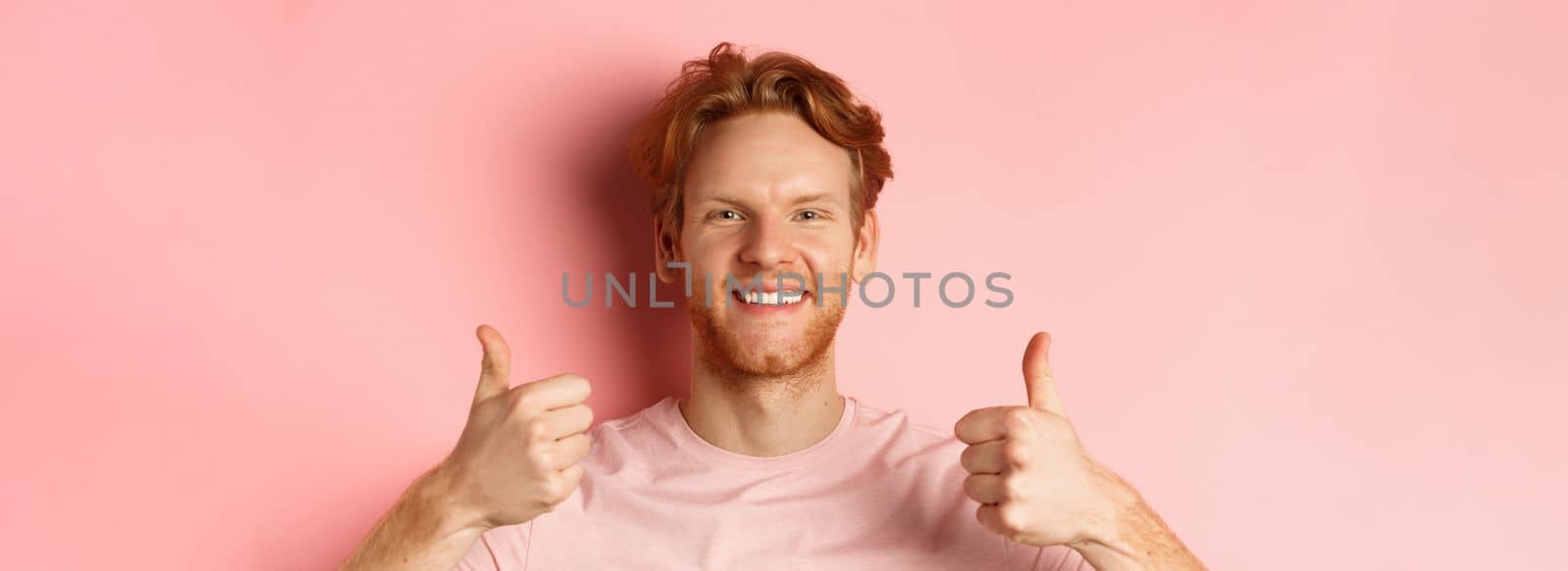 Close up of cheerful man with red hair and beard, showing thumbs up and smiling, saying yes, approve and praise something cool, standing over pink background.