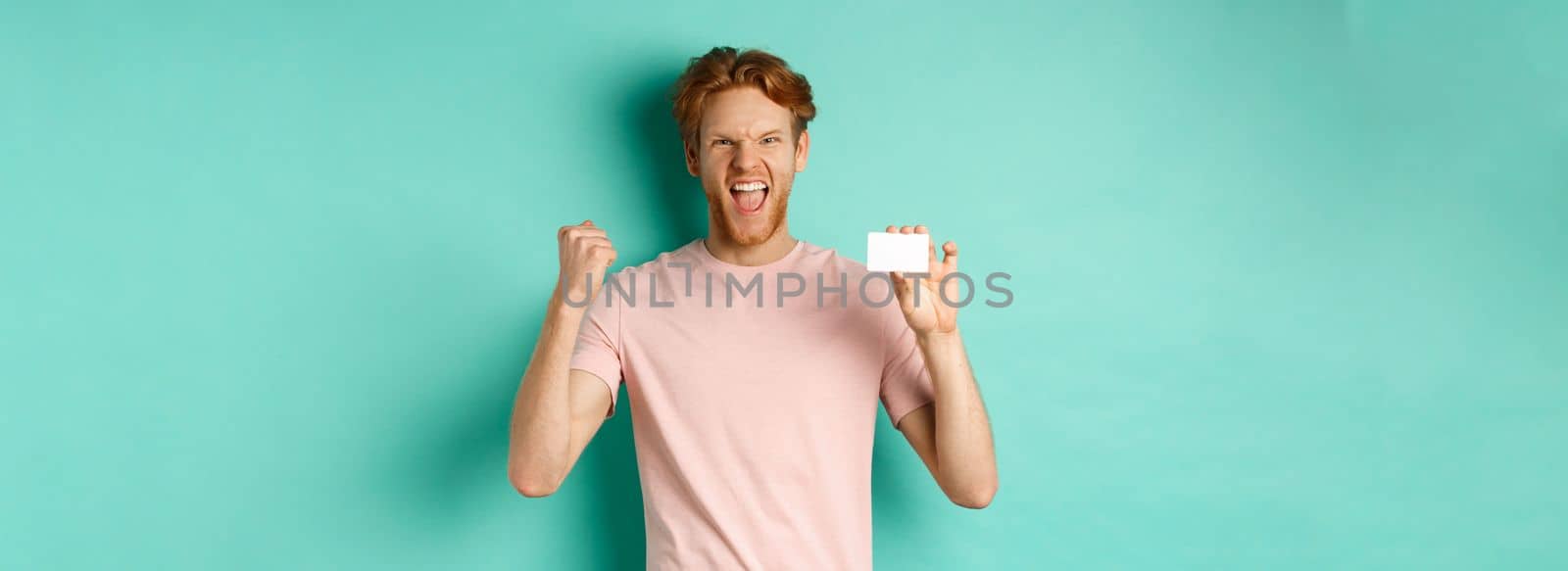 Cheerful young man triumphing, making fist pump to celebrate success, showing plastic credit card, winning prize from bank, turquoise background.