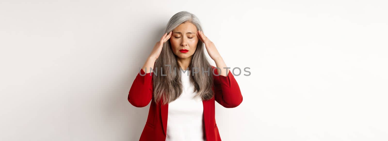 Distressed asian businesswoman trying to calm down, massaging temples on head to soothe headache, standing over white background.