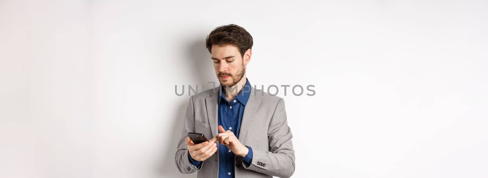 Handsome businessman in stylish suit chatting on smartphone, using mobile phone, standing on white background.