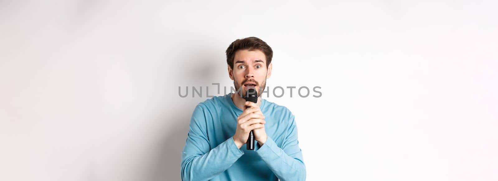 Confused young man looking nervously at camera while singing karaoke, holding microphone, standing over white background by Benzoix