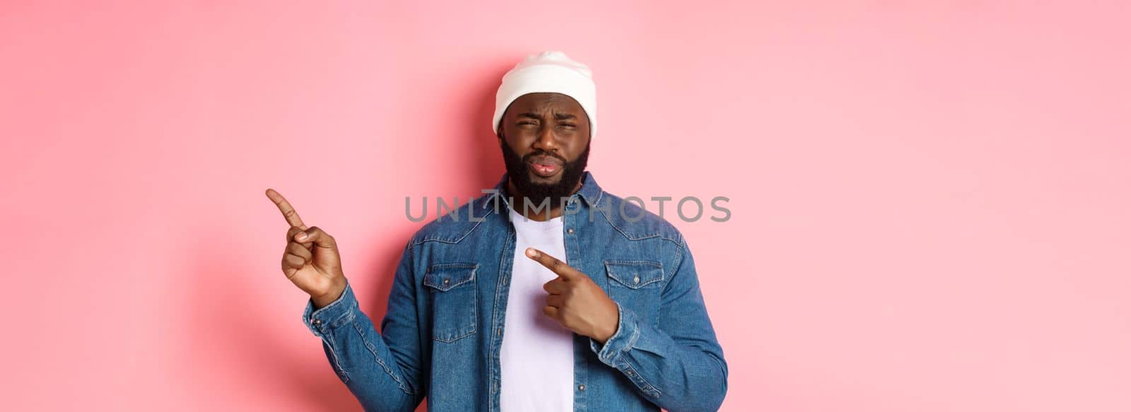 Disappointed and skeptical african-american guy complaining, cringe from disgust, frowning and grimacing from dissatisfaction, pointing fingers right, pink background.