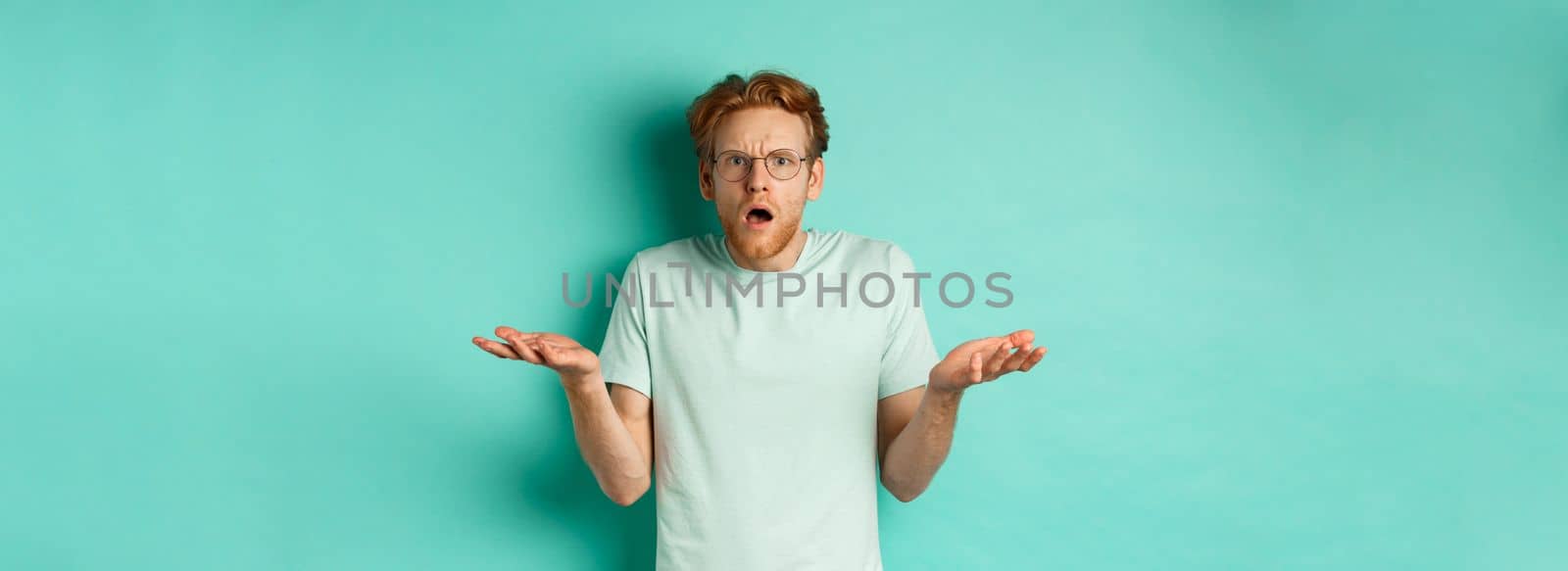 Confused and shocked guy with red messy hair and glasses, shrugging and raising hands, staring at something strange, cant understand, standing over turquoise background.