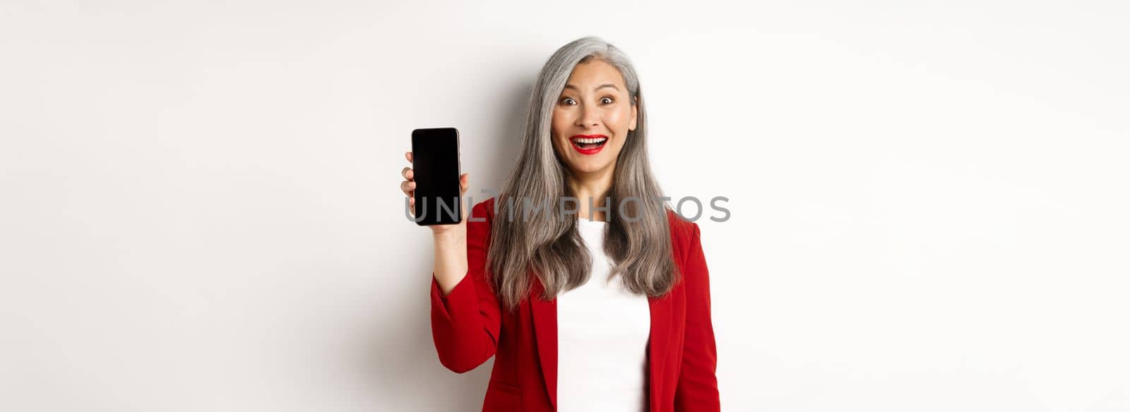 Cheerful asian female entrepreneur showing mobile phone screen, smiling amused, standing over white background by Benzoix
