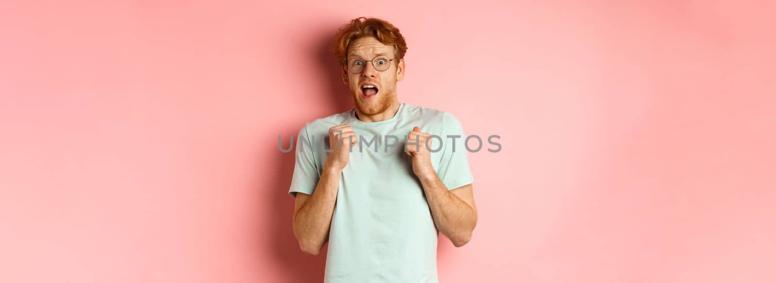 Portrait of scared redhead guy in glasses staring startled at camera, press hands to body and scream of fear, standing over pink background.