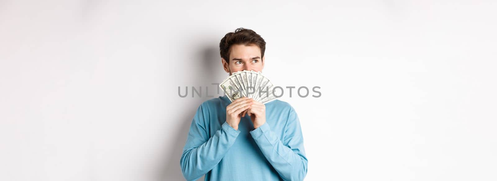 Dreamy man thinking of buying product, holding money and looking left pensive, imaging shopping, standing over white background.