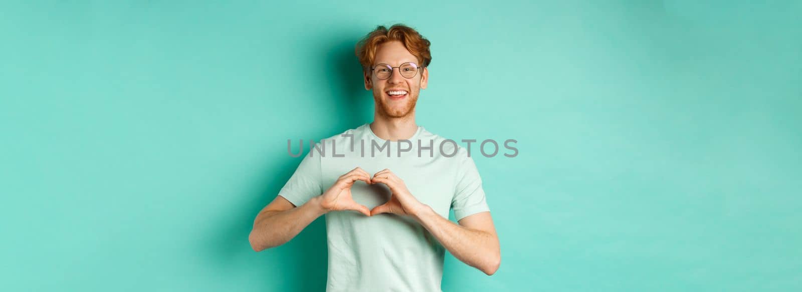 Valentines day and relationship concept. Happy boyfriend with red hair and beard, wearing glasses and t-shirt, showing heart sign and saying I love you, standing over turquoise background.