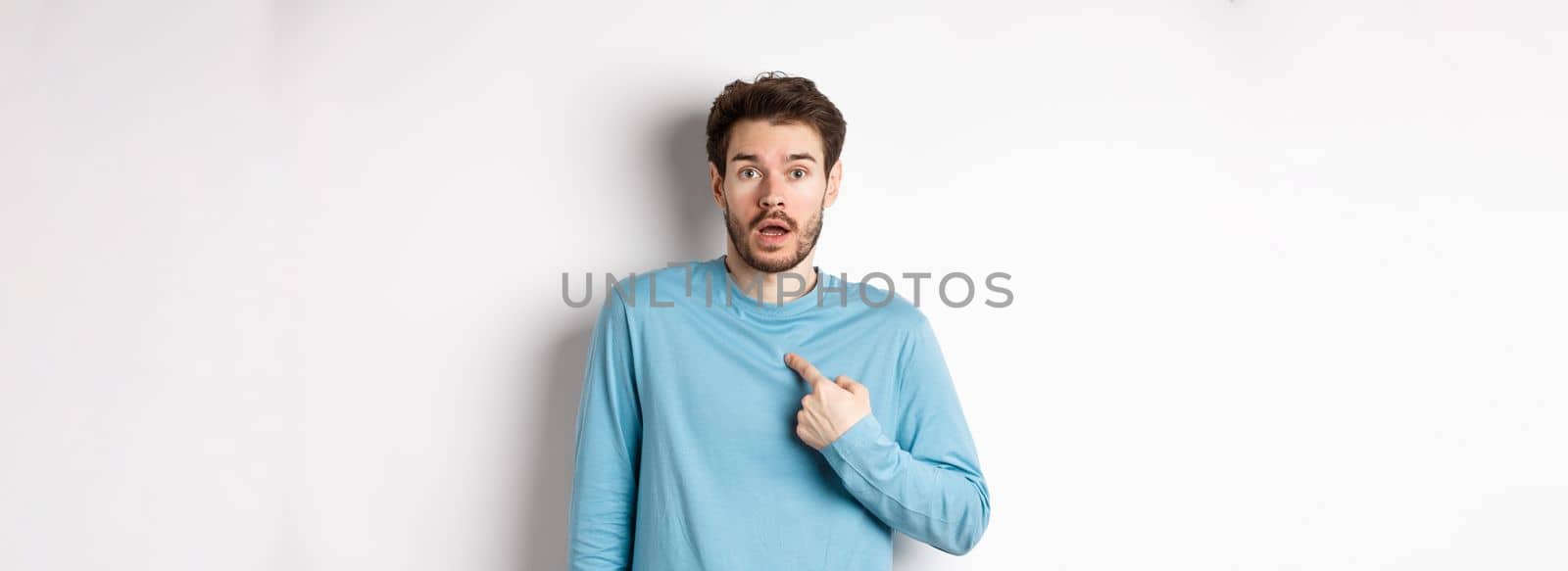 Surprised young man gasping and pointing at himself with disbelief, being chosen or named, standing over white background.