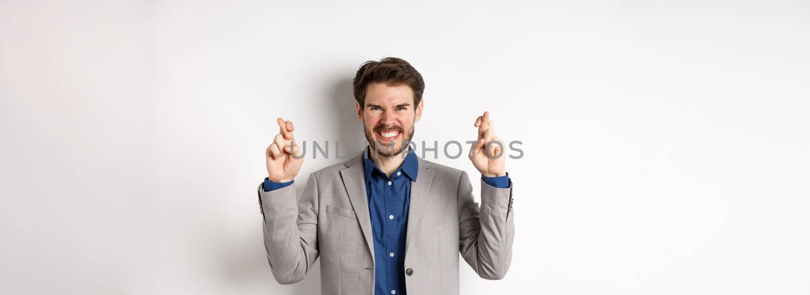 Motivated and excited businessman smiling positive, look hopeful and cross fingers for good luck, making wish, waiting for announcement, white background by Benzoix