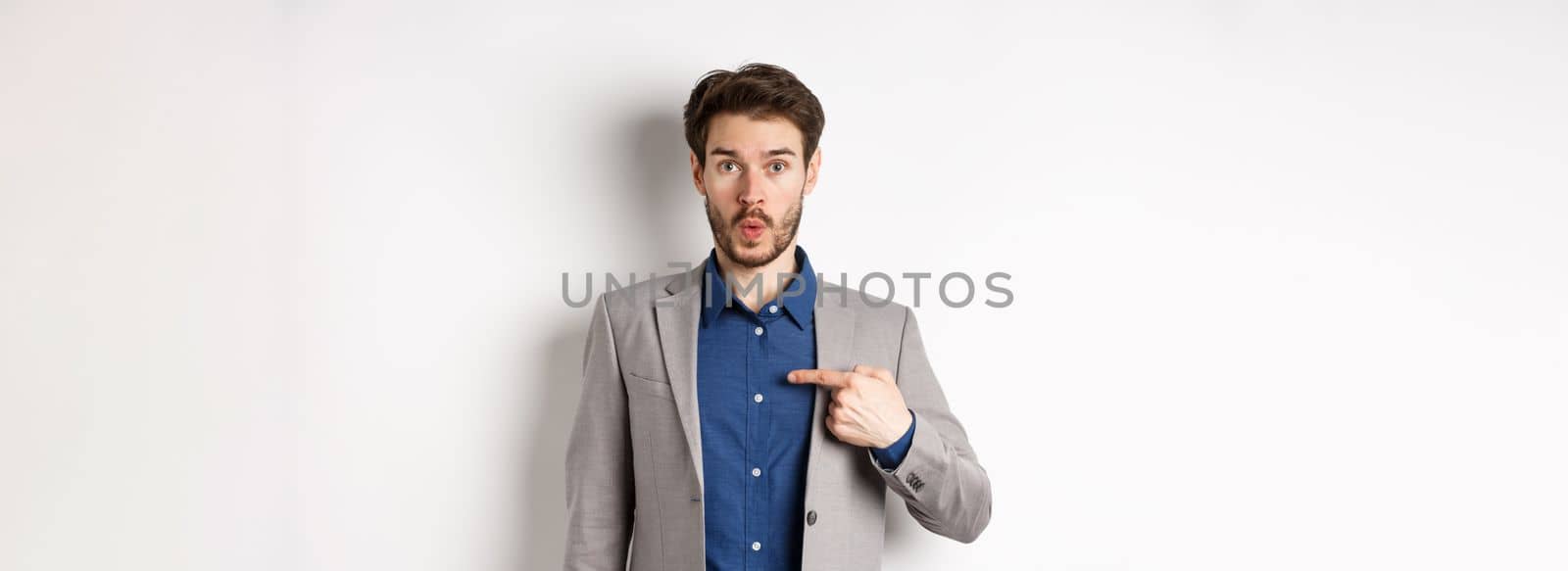 Who me. Surprised man in suit pointing at himself with disbelief, looking confused at camera, standing on white background.