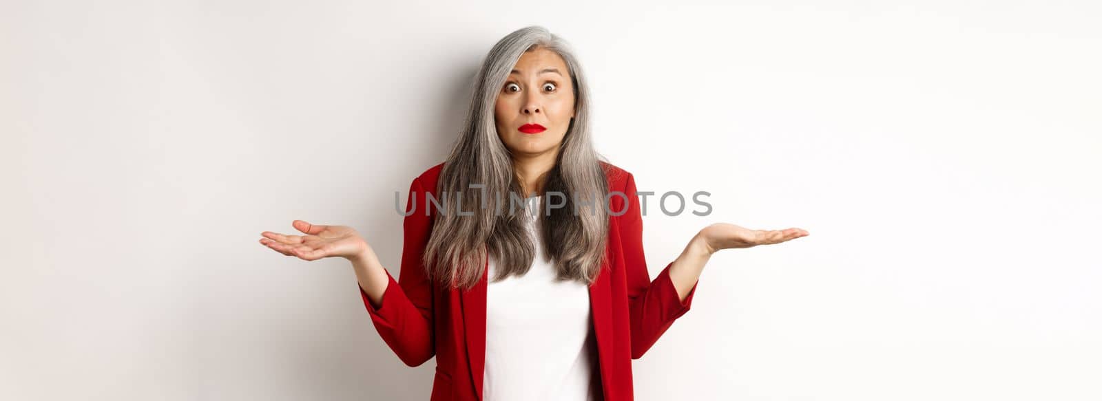 Confused asian senior woman shrugging, spread hands sideways and staring questioned at camera, dont know anything, standing over white background by Benzoix