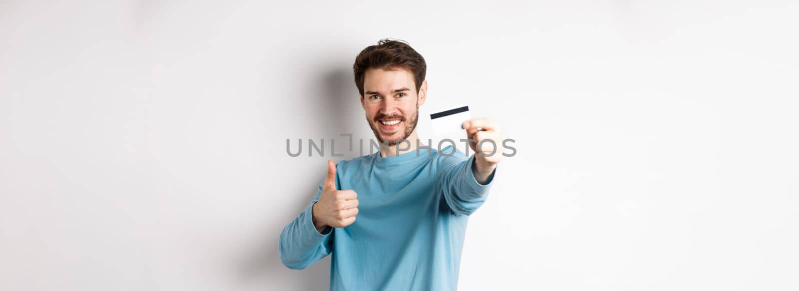Handsome caucasian man showing plastic credit card with thumb-up, recommend and praise good bank offer, standing on white background