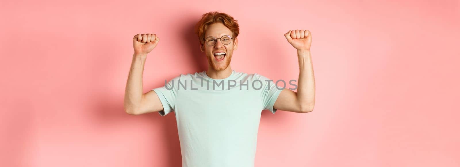 Happy winner with red hair and beard celebrating victory, shouting yes with joy and raising hands up, enjoy success, standing over pink background.