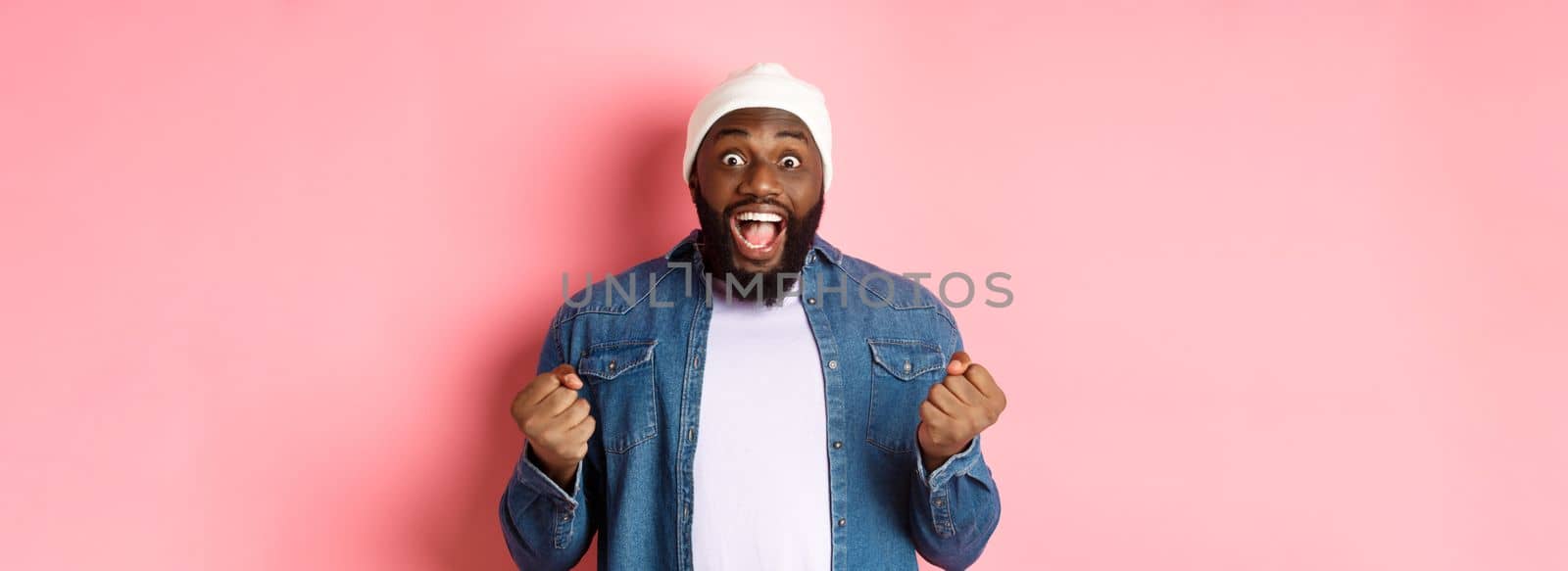 Surprised and happy man staring with disbelief while winning, clench fists and smiling amazed, celebrating victory, achieve goal, standing over pink background by Benzoix