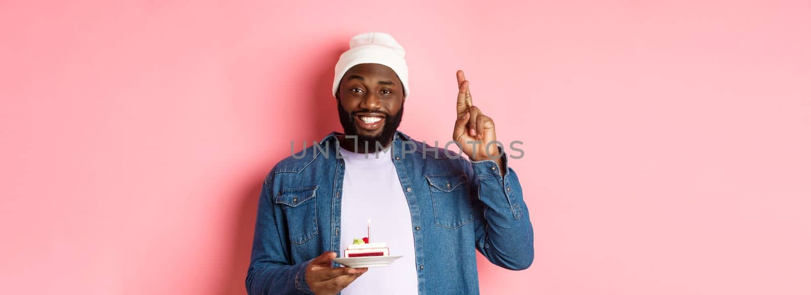 Handsome african-american guy celebrating birthday, making wish with fingers crossed, holding bday cake with candle, standing against pink background by Benzoix