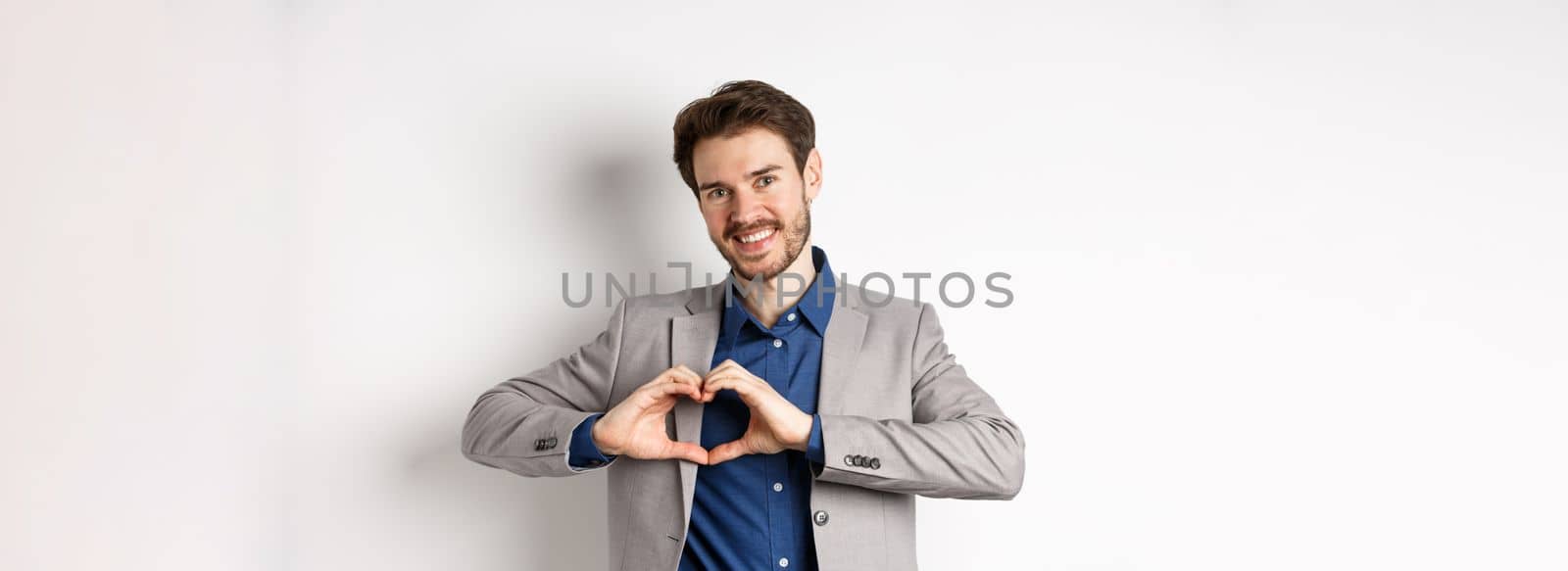 Romantic man in suit showing heart sign and smiling, love his girlfriend, standing on white background, making confession to lover.