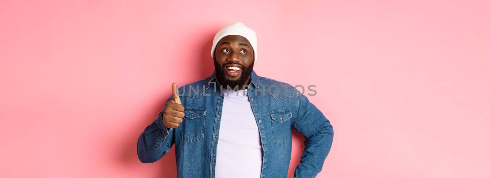 Happy Black man showing thumb-up, looking at upper left corner thoughtful, standing over pink background by Benzoix