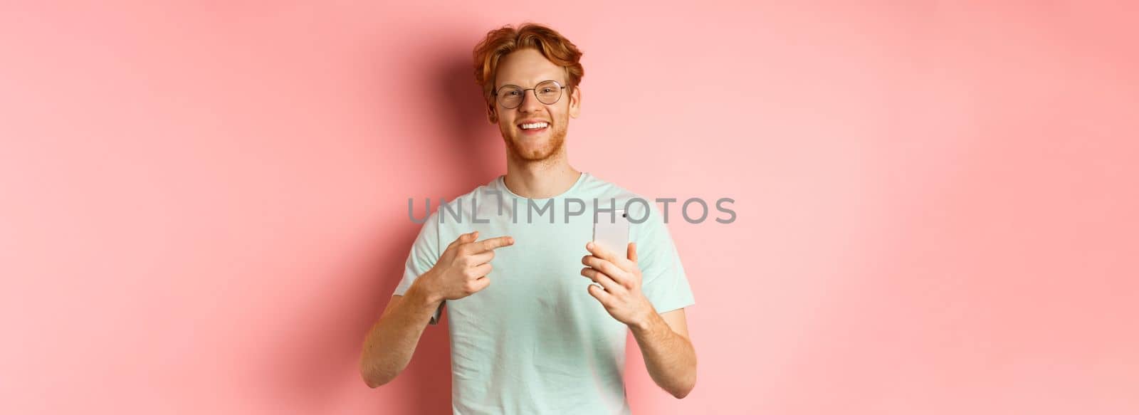 Young man with red hair and beard, wearing t-shirt and glasses, smiling while pointing finger at smartphone, recommend online promotion, pink background by Benzoix