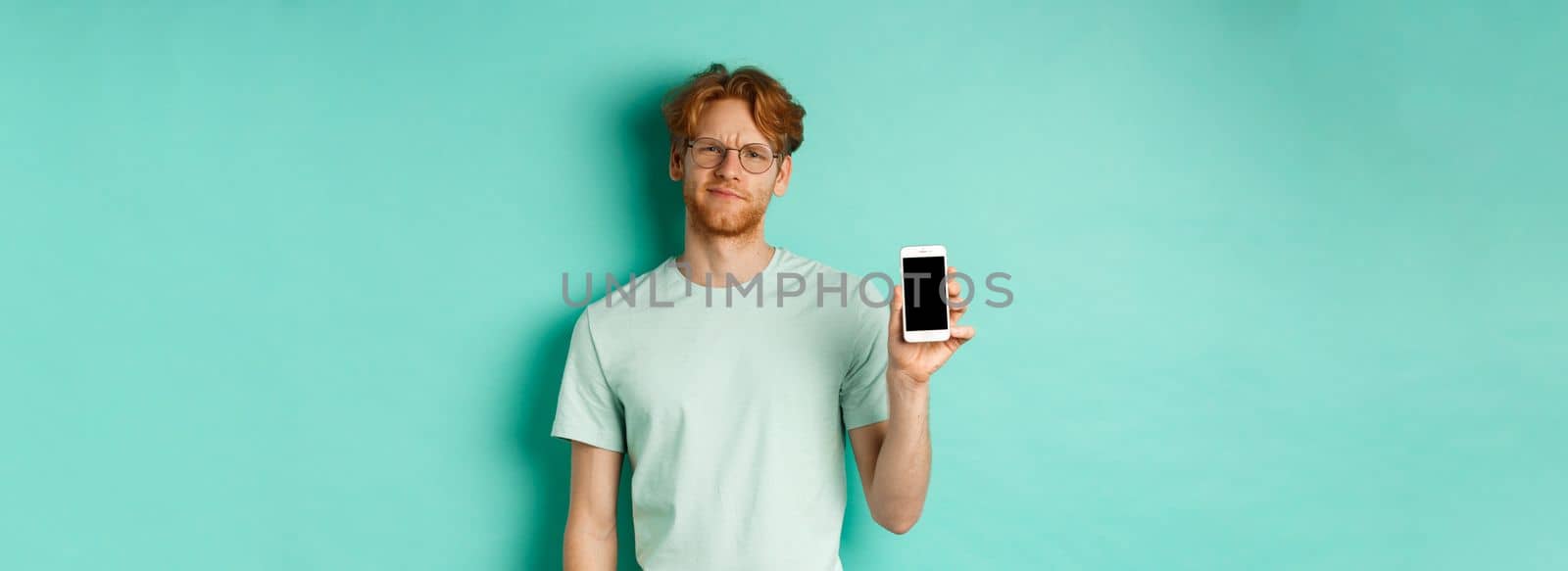 Skeptical male model with red hair and glasses, showing mobile screen and frowning disappointed, dislike application, standing over turquoise background.