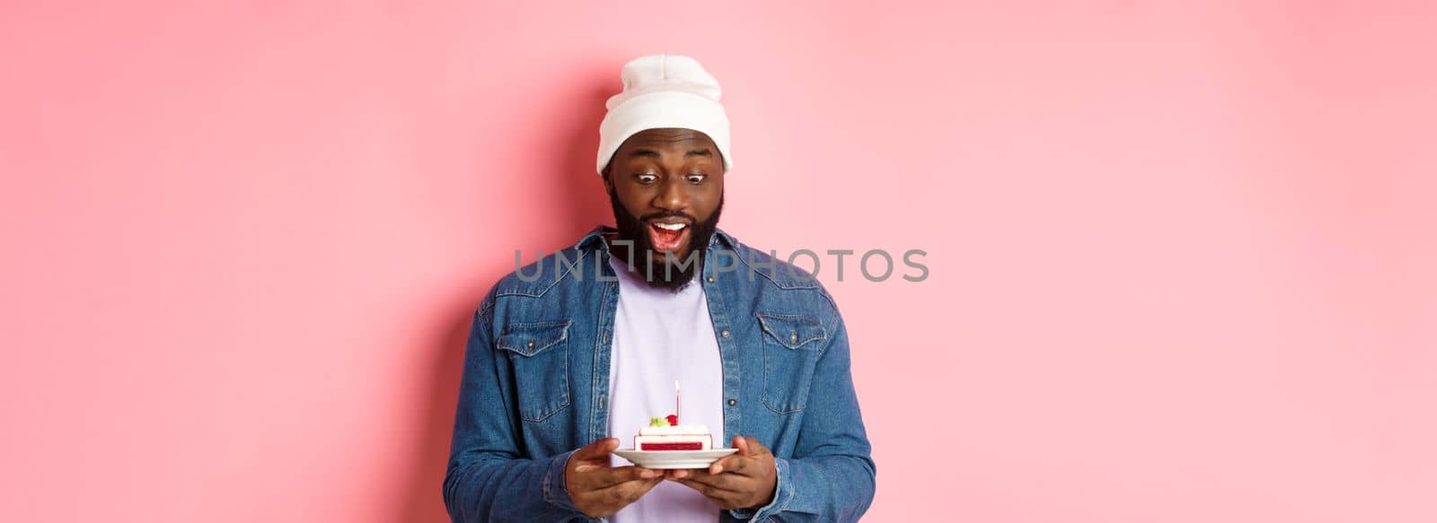 Cheerful african-american guy celebrating birthday, making wish on bday cake with lit candle, smiling happy, standing over pink background.