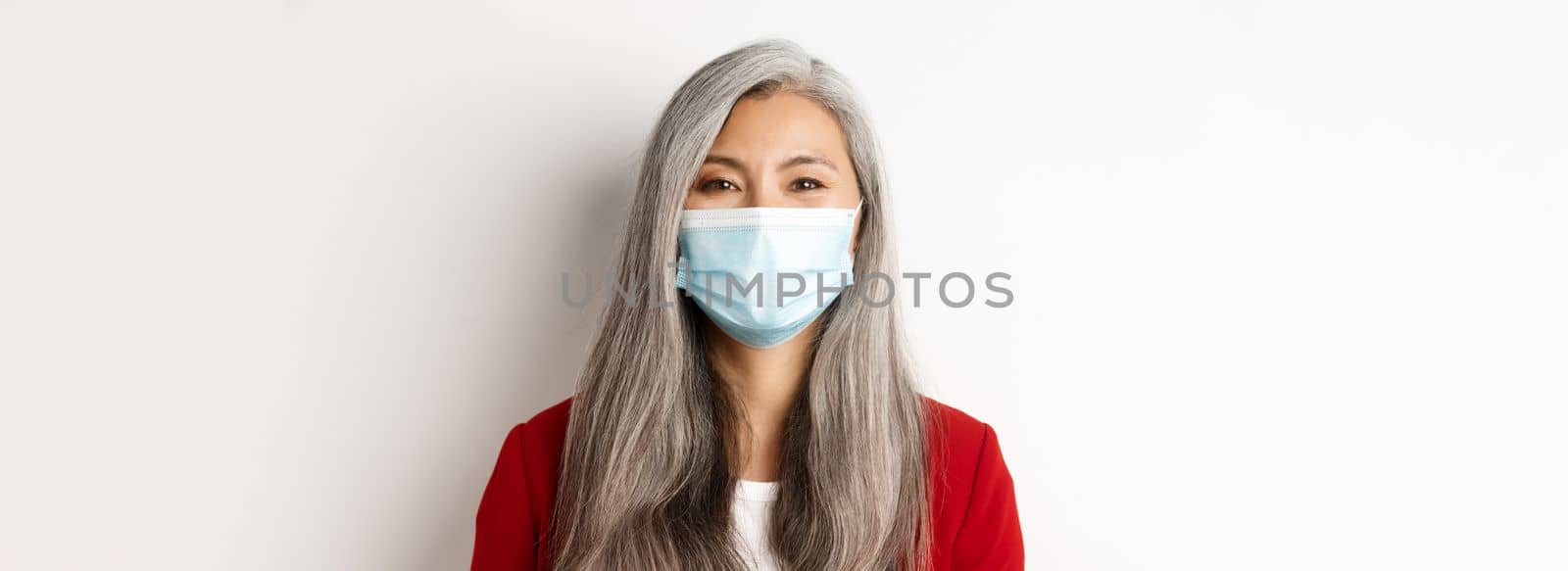 Covid, pandemic and business concept. Close-up of cheerful asian senior businesswoman in medical mask smiling at camera, white background.