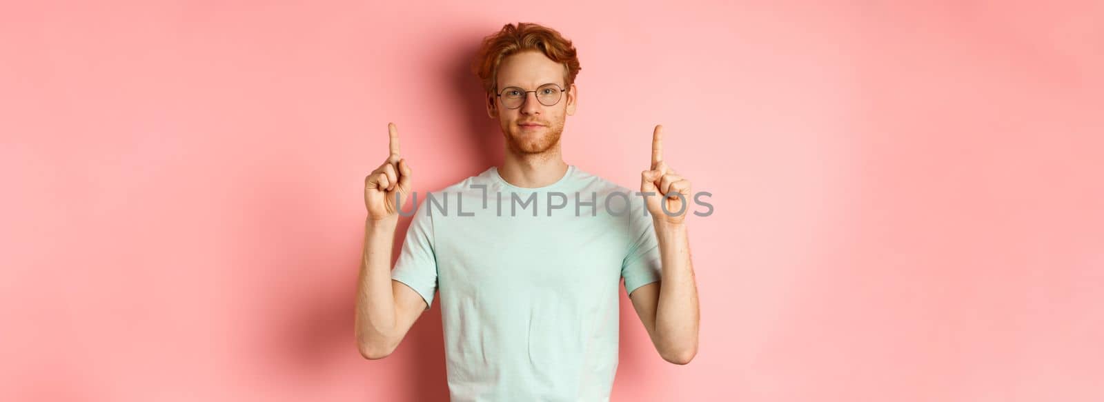 Confident and pleased young man with red hair, wearing glasses and t-shirt, pointing fingers up and smiling with smug face, showing good deal, standing over pink background.