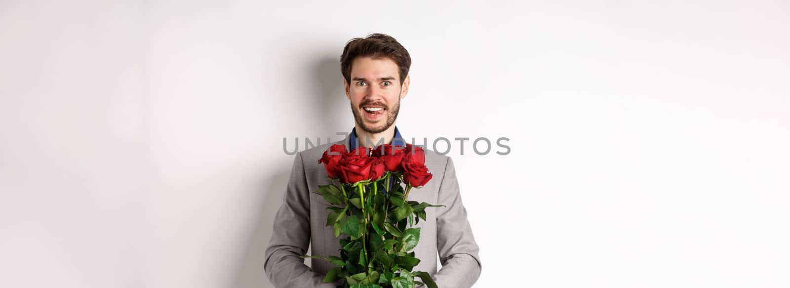 Excited handsome man in suit holding bouquet of roses for romantic date with lover, standing happy on Valentines day, white background by Benzoix