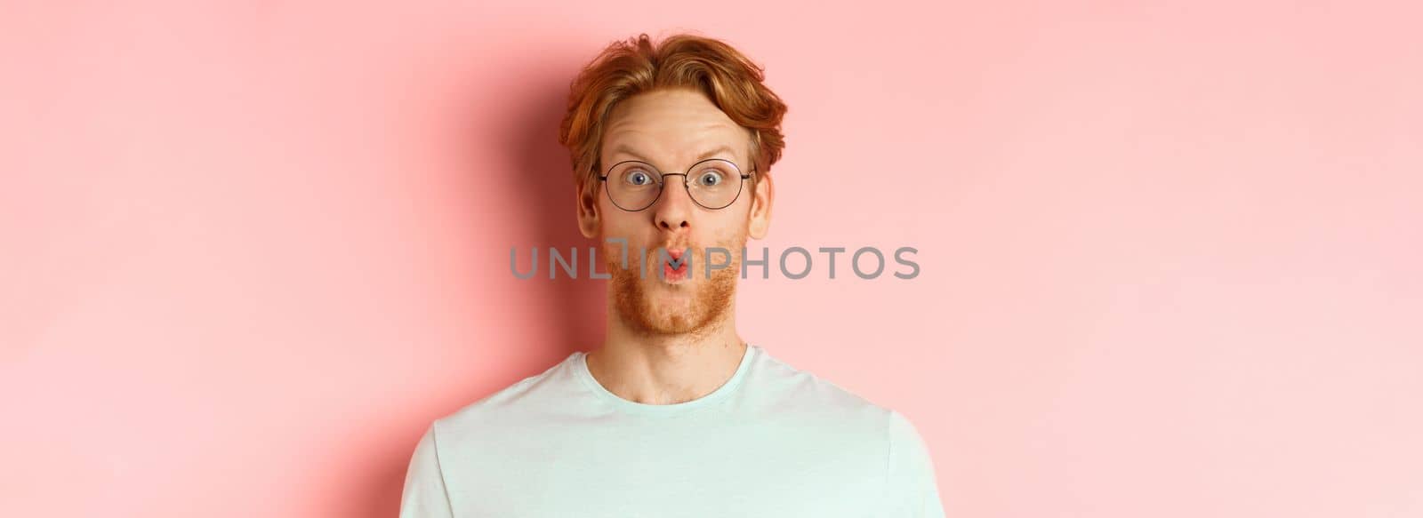 Close up of impressed redhead man in glasses saying wow, raising eyebrows surprised and staring at camera, standing over pink background.